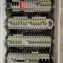 Rozdzielnica elektryczna z zamontowanymi bezpiecznikami, okablowaniem i wyświetlaczem wskazującym napięcie. Widoczne elementy instalacji elektrycznej wewnątrz obudowy.