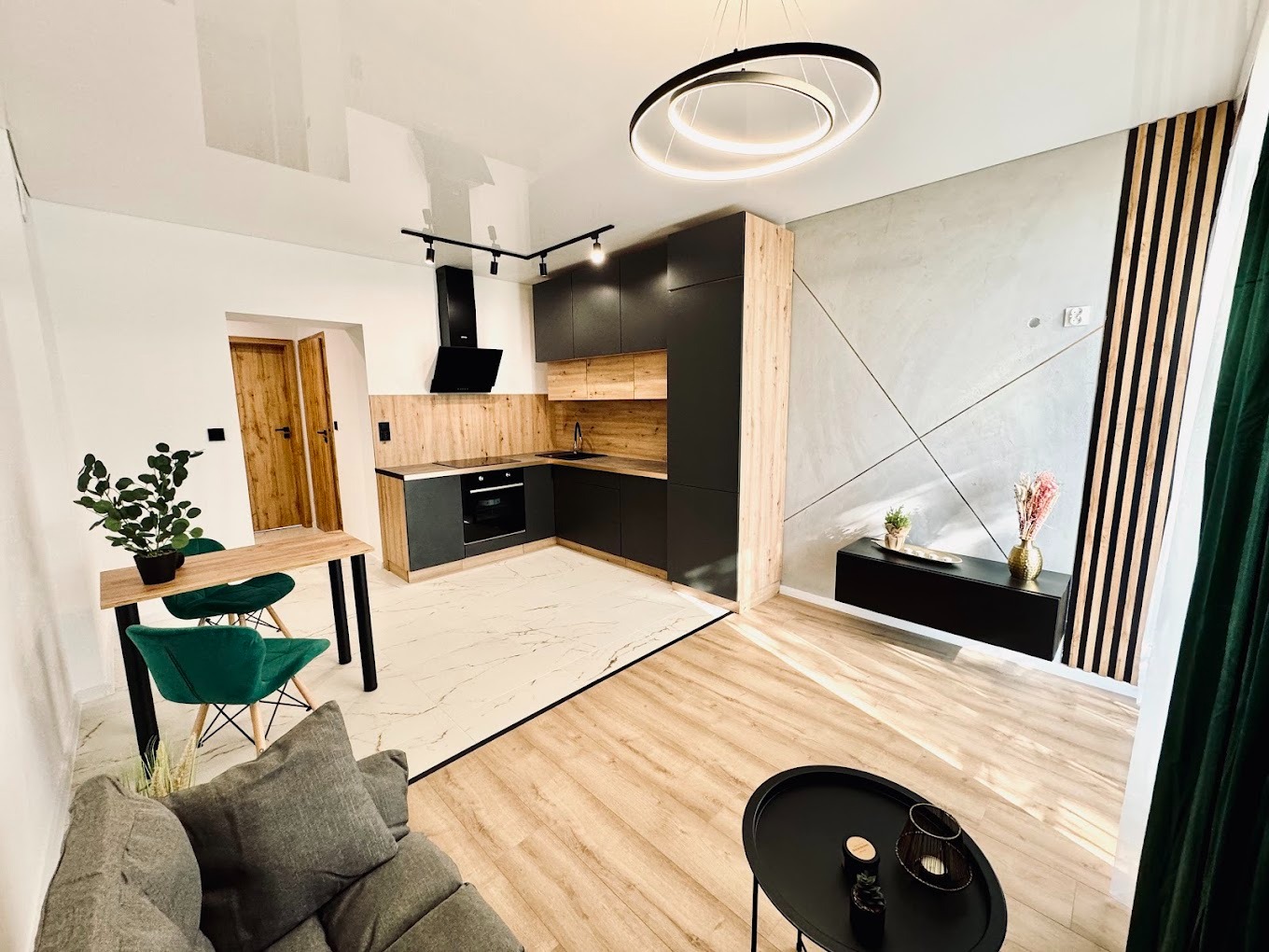 Nowoczesna kuchnia z czarnymi szafkami, drewnianymi akcentami i okrągłą lampą sufitową. Widoczny fragment salonu z szarą sofą i stolikiem.