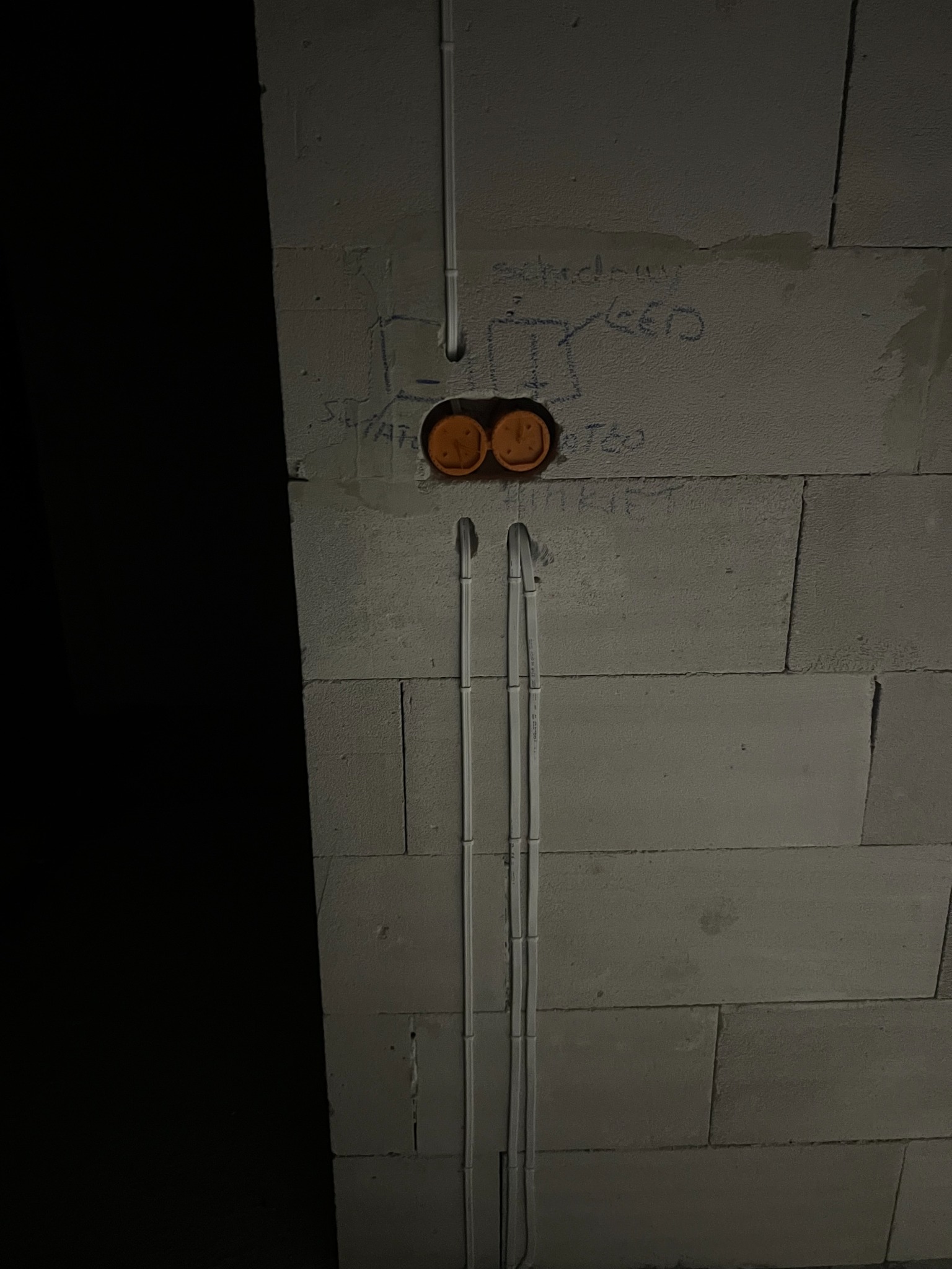 Szara ściana z betonu komórkowego z przewodami elektrycznymi w peszlach, puszką podwójną i schematem instalacji narysowanym na ścianie. Etap prac instalacyjnych.