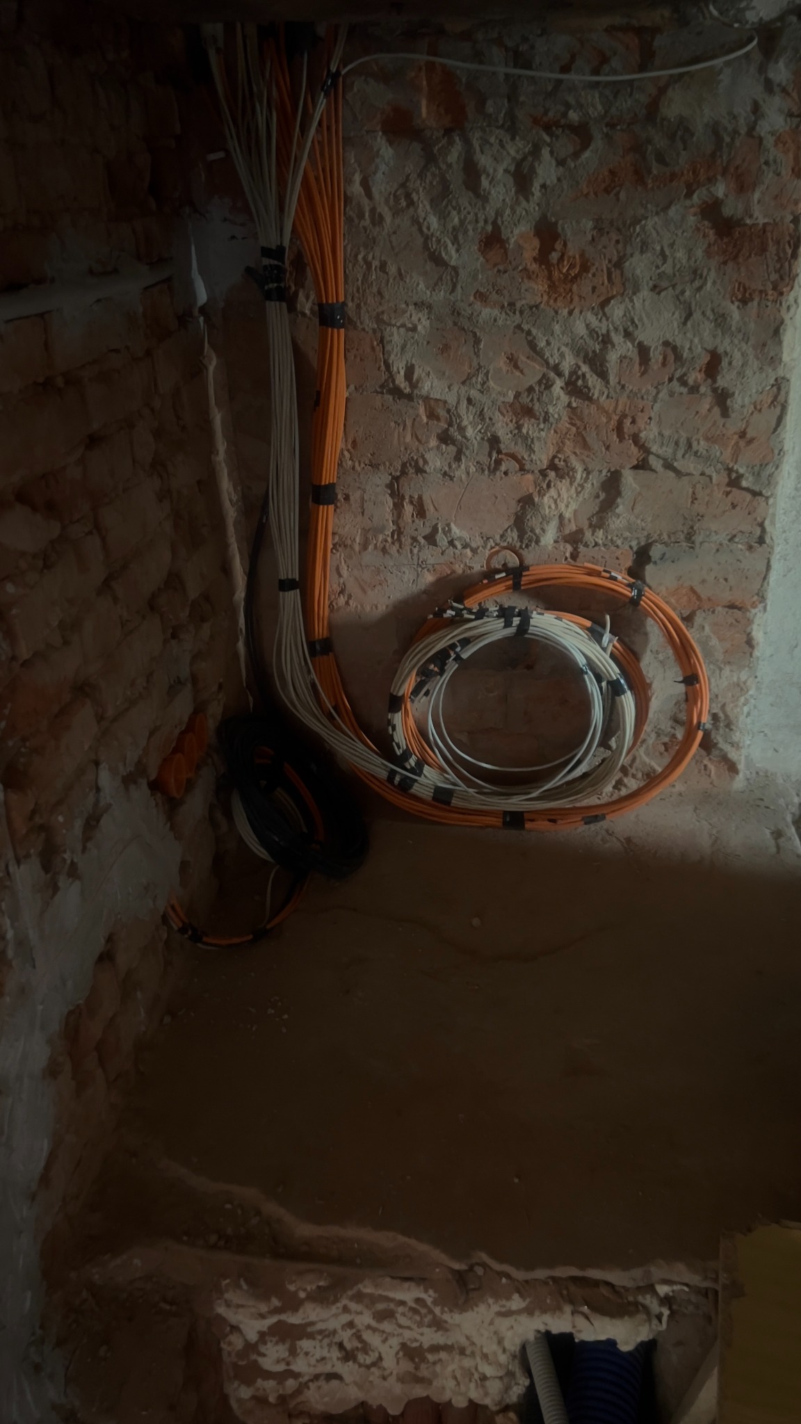 Kłębek pomarańczowych i białych kabli elektrycznych, spiętych czarną taśmą, zwisających ze ściany z cegły w surowym stanie. Widoczne peszle instalacyjne.