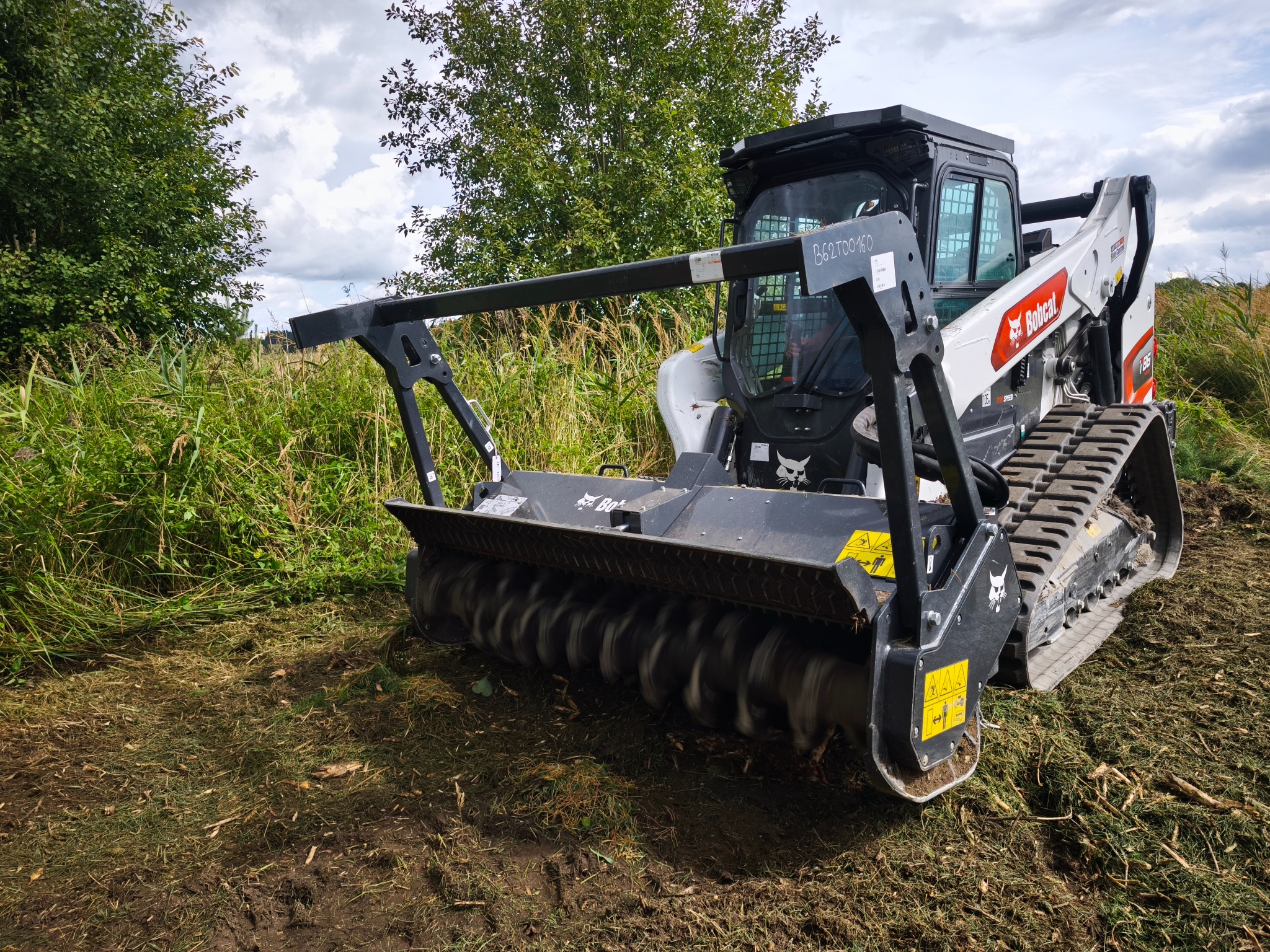 Bobcat z frezem leśnym w trakcie karczowania terenu, widoczne resztki po wycince. Tło: drzewa i pochmurne niebo. Maszyna w akcji, świeżo skoszona trawa.