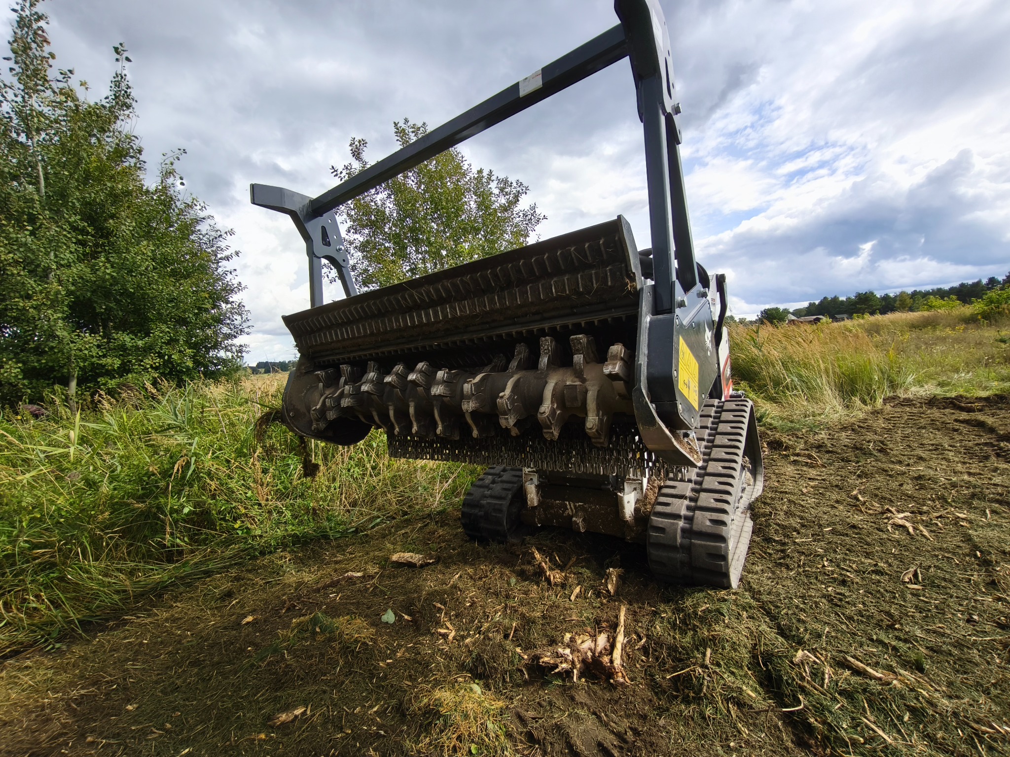 Szerokokątne ujęcie mulczera gąsienicowego w trakcie pracy na zarośniętym terenie. Świeżo zmielony materiał roślinny widoczny na ziemi, drzewa w tle.