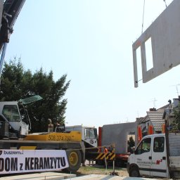 BUSZREM SPÓŁKA AKCYJNA - Montaż prefabrykowanego elementu ściany z otworem okiennym za pomocą dźwigu na placu budowy, widoczny baner reklamowy z nazwą firmy. W tle zabudowa jednorodzinna.