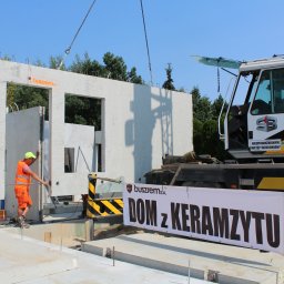 BUSZREM SPÓŁKA AKCYJNA - Montaż prefabrykowanych ścian z keramzytu na budowie domu jednorodzinnego przy użyciu dźwigu; pracownicy w kamizelkach odblaskowych i kaskach stabilizują element, w tle baner reklamowy 'Dom...