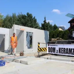 Montaż prefabrykowanych ścian na budowie domu jednorodzinnego; pracownicy w pomarańczowych kamizelkach, widoczny dźwig z banerem reklamowym.