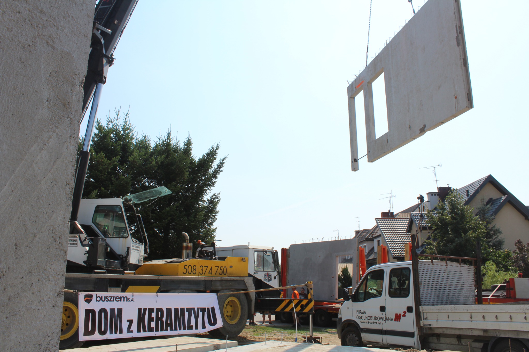 Montaż prefabrykowanego elementu ściany z otworem okiennym za pomocą dźwigu na placu budowy, widoczny baner reklamowy z nazwą firmy. W tle zabudowa jednorodzinna.