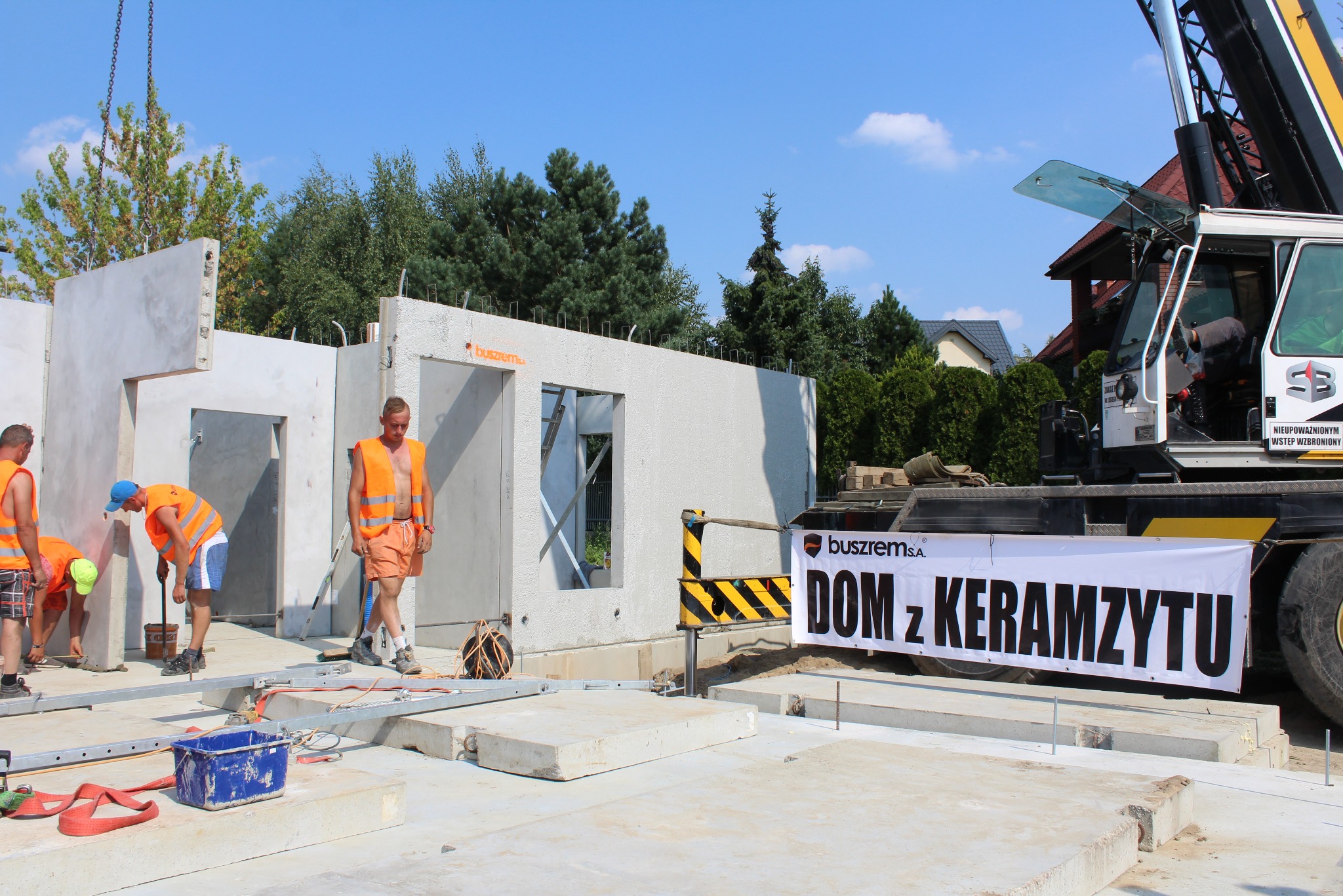 Montaż prefabrykowanych ścian na budowie domu jednorodzinnego; pracownicy w pomarańczowych kamizelkach, widoczny dźwig z banerem reklamowym.