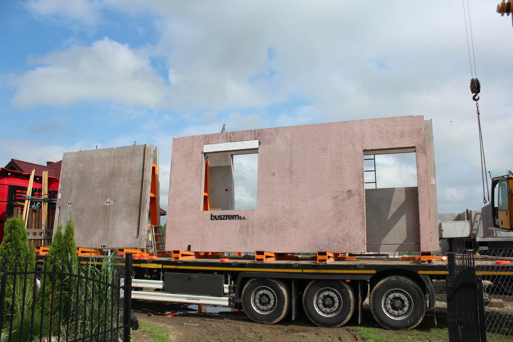 Transport prefabrykowanych ścian z otworami okiennymi na naczepie ciężarówki, widoczny dźwig i fragment budynku w tle.