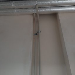 HYDROLUD ŁUKASZ LODZIŃSKI - Dwie pionowe, białe rury przymocowane do ściany za pomocą metalowych uchwytów, z widocznymi poziomymi rurami w izolacji na suficie.