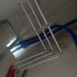 HYDROLUD ŁUKASZ LODZIŃSKI - Szczegół instalacji rur: białe rury przymocowane do sufitu za pomocą metalowych uchwytów, obok niebieskich i czerwonych rur, widoczne elementy konstrukcji sufitu podwieszanego.