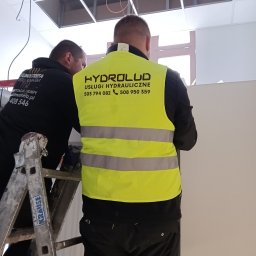 HYDROLUD ŁUKASZ LODZIŃSKI - Dwóch hydraulików w kamizelkach odblaskowych z logo firmy, wykonuje prace instalacyjne w pomieszczeniu z podwieszanym sufitem.