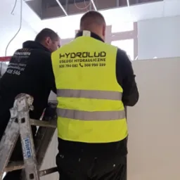 Dwóch hydraulików w kamizelkach odblaskowych z logo firmy, wykonuje prace instalacyjne w pomieszczeniu z podwieszanym sufitem.