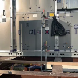 Duża centrala klimatyzacyjna na dachu budynku, widoczne liczne panele z oznaczeniami, rury i zawory, oraz elementy konstrukcji wsporczej.