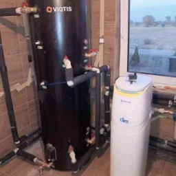 Czarny zasobnik ciepłej wody użytkowej Viqtis z podłączeniami i biały zmiękczacz wody Klarwod w pomieszczeniu z oknem i brązowymi płytkami.