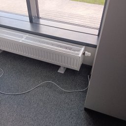 HYDROLUD ŁUKASZ LODZIŃSKI - Biały, żebrowany grzejnik panelowy zamontowany pod oknem na szarym dywanie. Widoczny termostat i kabel. Ujęcie z niskiego kąta, z boku.