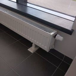 HYDROLUD ŁUKASZ LODZIŃSKI - Biały grzejnik panelowy z termostatem, zamontowany pod oknem na ciemnej, kwadratowej płytce podłogowej. Widok z boku, z zewnątrz budynku w tle.