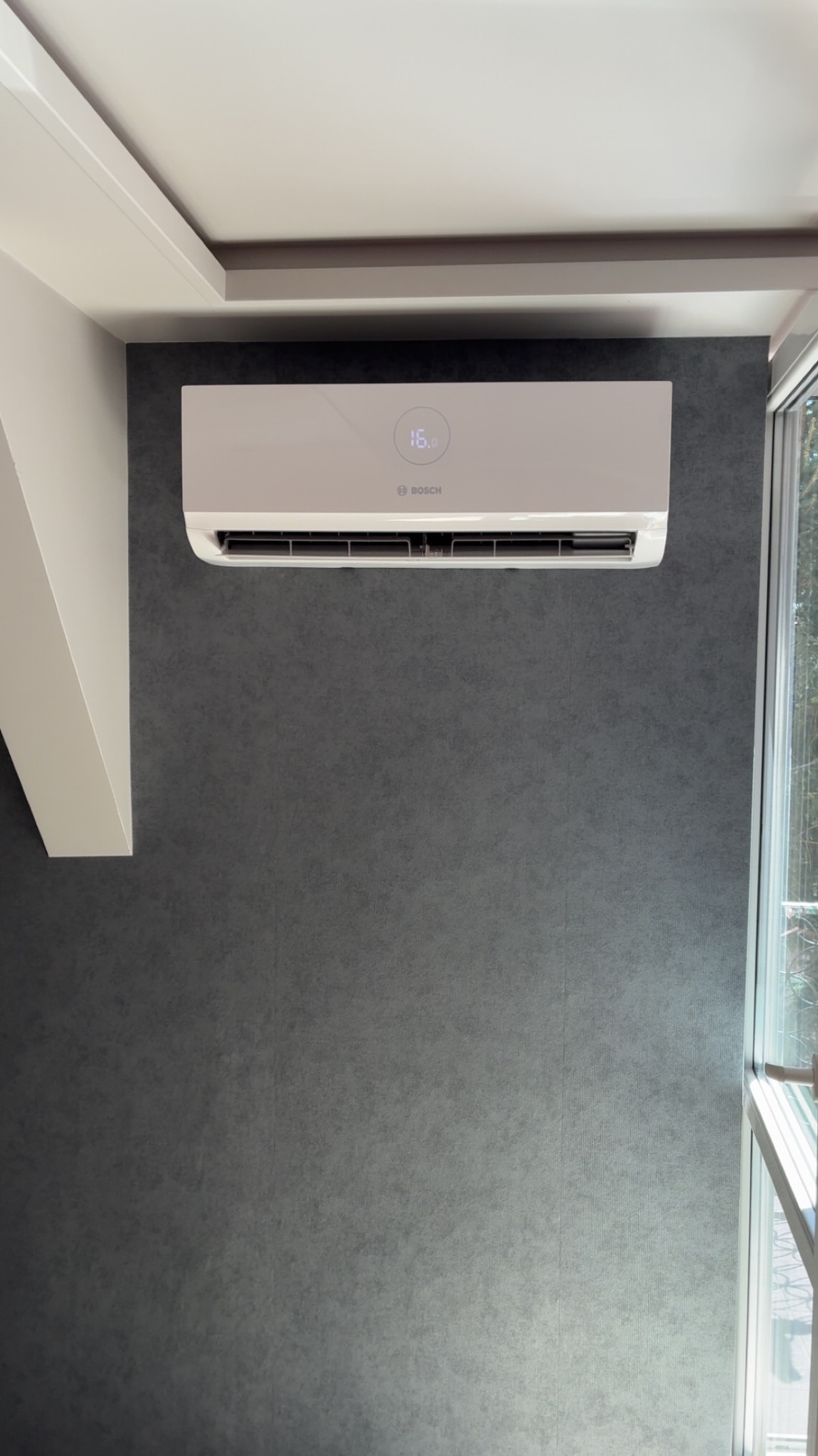 Biały klimatyzator Bosch na szarej ścianie z oknem. Urządzenie włączone, wyświetla temperaturę 16 stopni Celsjusza. Minimalistyczny design, nowoczesne wnętrze.