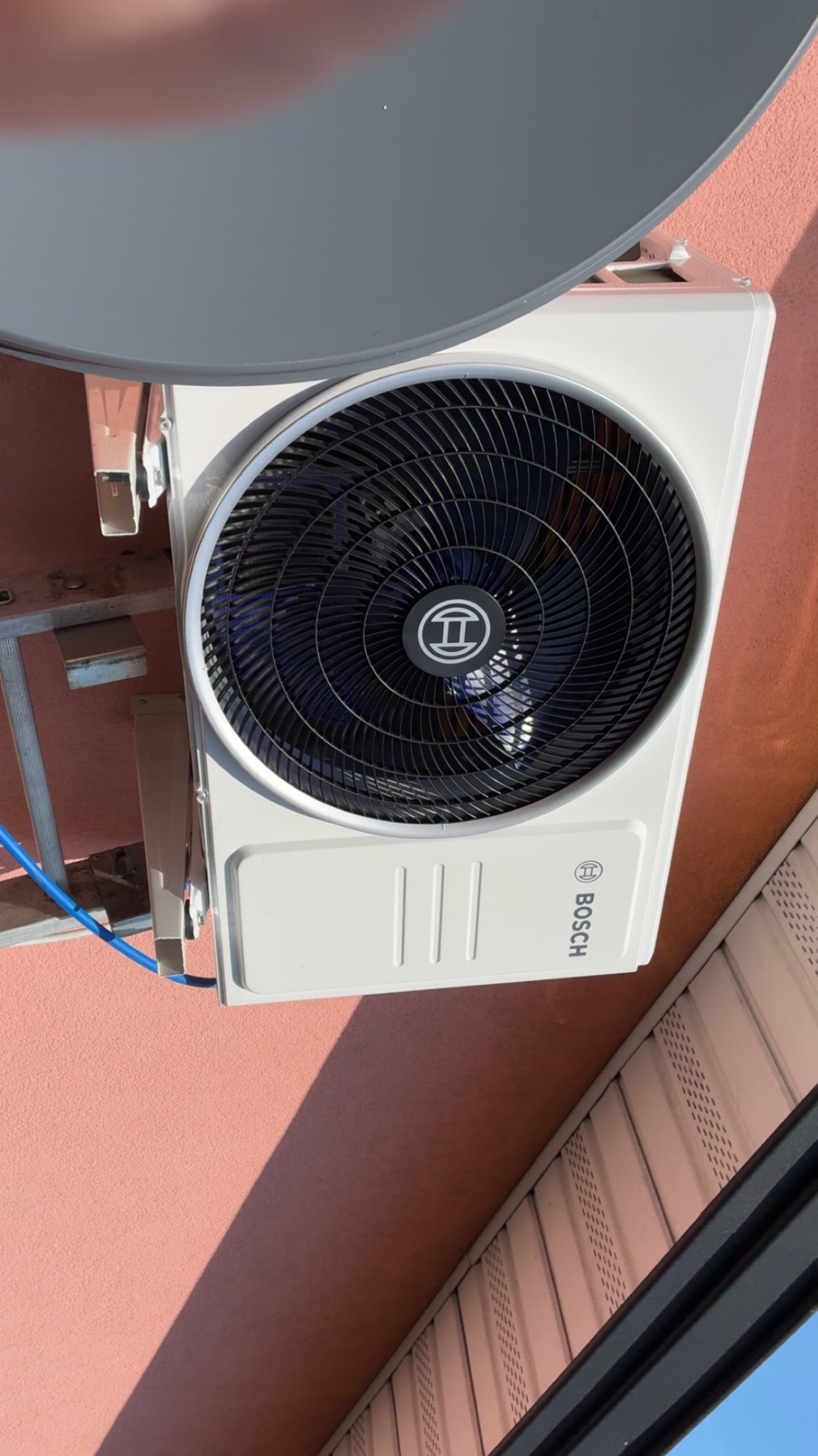 Biała jednostka zewnętrzna klimatyzacji Bosch, zamontowana na różowej ścianie budynku, z fragmentem szarej anteny satelitarnej w górnej części kadru. Widoczne elementy montażowe.