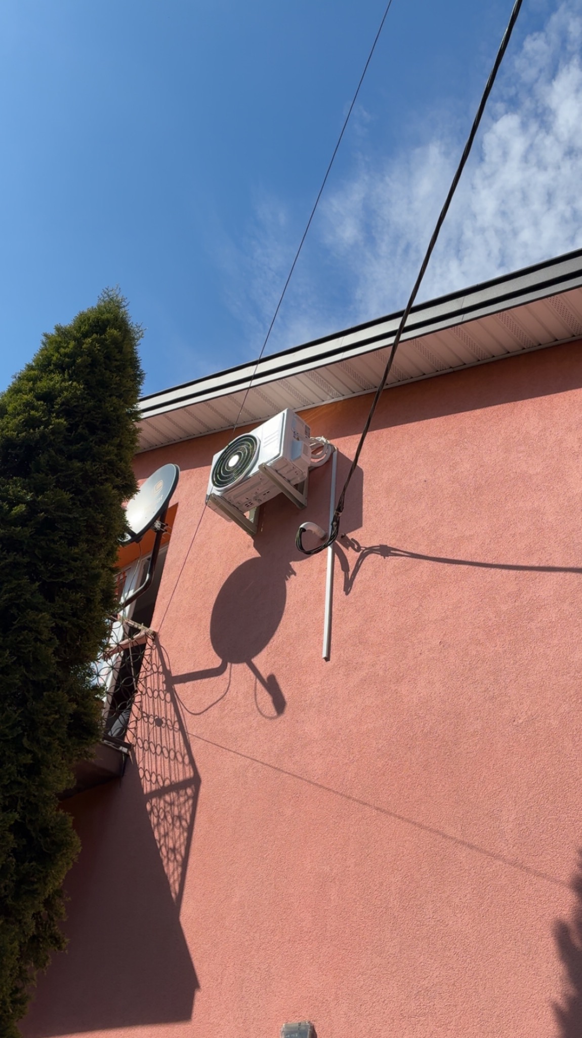 Biały klimatyzator zamontowany na różowej ścianie budynku, widoczne przewody, antena satelitarna i cień urządzenia. Błękitne niebo w tle.