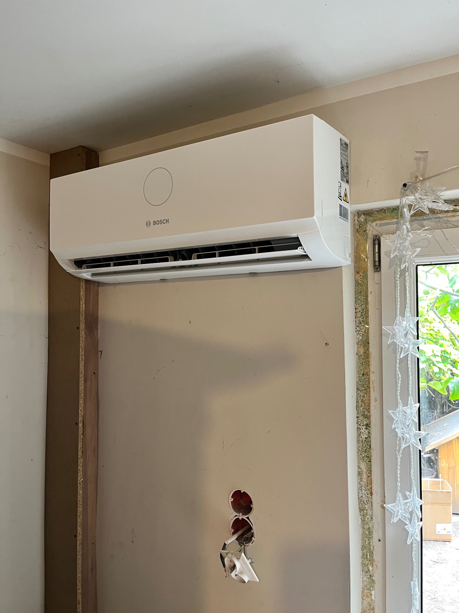 Zamontowana klimatyzacja Bosch na ścianie w trakcie remontu, widoczne niedokończone wykończenia i otwory instalacyjne. Oświetlenie naturalne, dekoracje okienne.