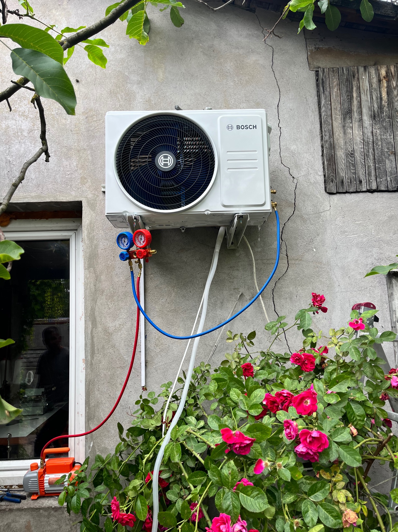 Biały klimatyzator Bosch na szarej ścianie, podłączony do manometrów i pompy próżniowej, otoczony zielenią i różami. Widoczna instalacja w trakcie serwisu.