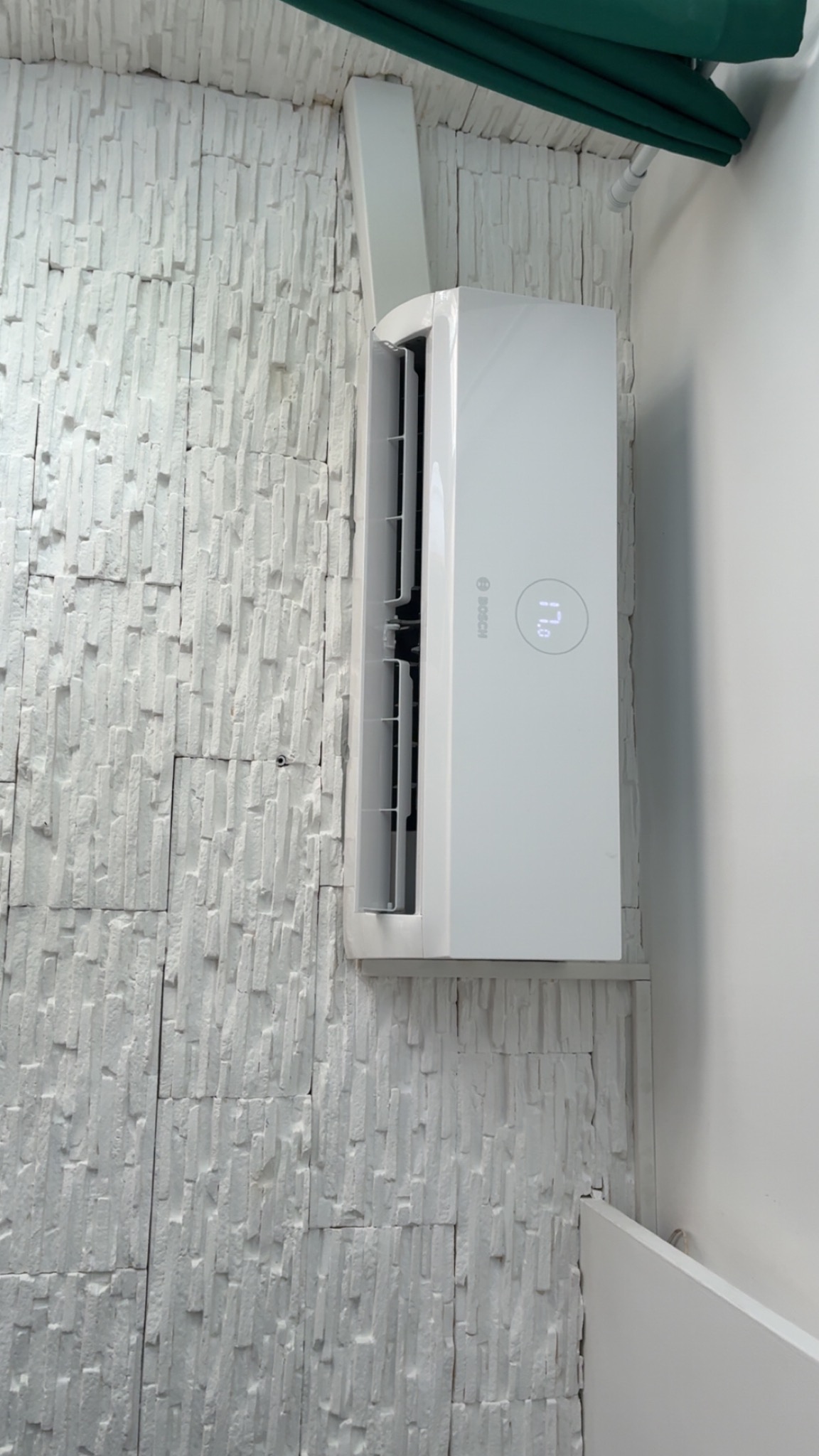 Biały klimatyzator Bosch na ścianie z dekoracyjnej, białej cegły. Widoczny fragment zielonego karniszu. Estetyczny montaż, nowoczesny design.