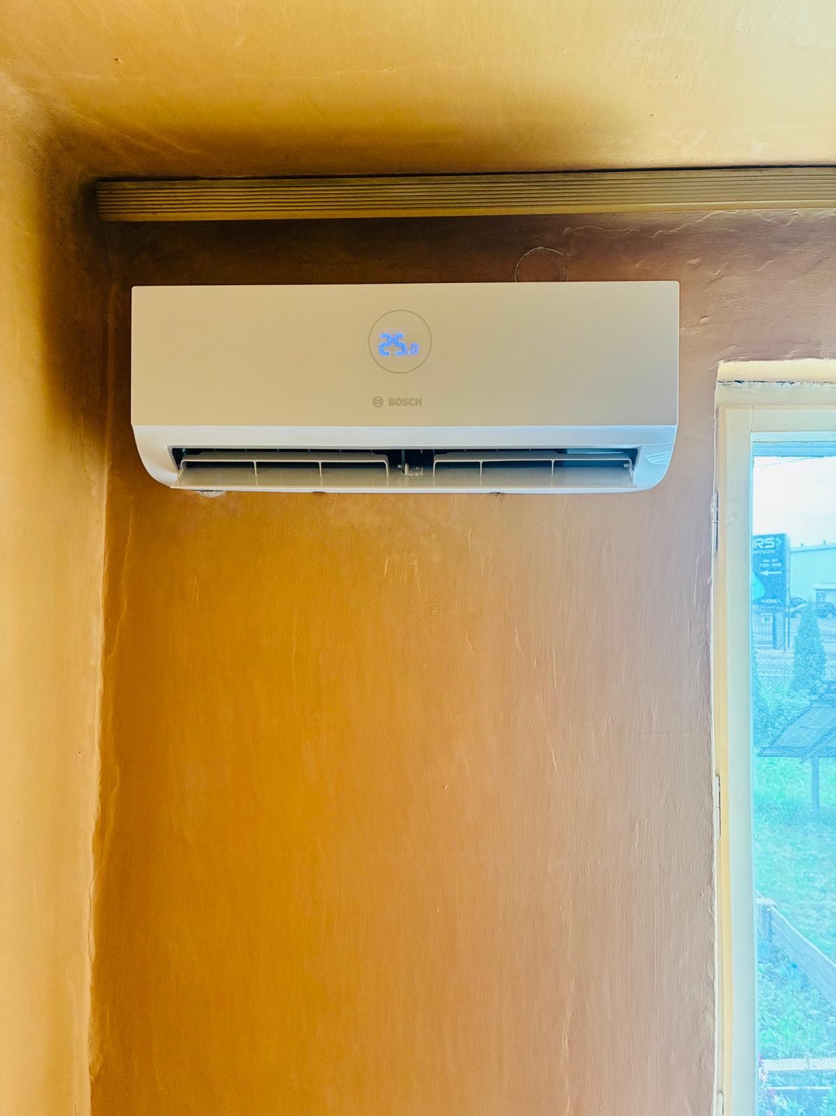 Biały klimatyzator Bosch zamontowany na ścianie w pomieszczeniu. Na wyświetlaczu widoczna temperatura. Obok okno z widokiem na zewnątrz.