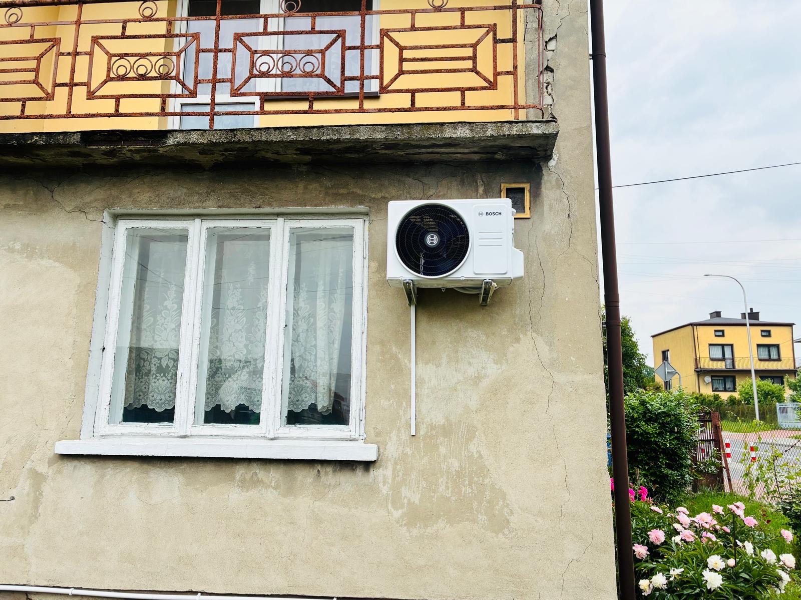 Biały klimatyzator Bosch zamontowany na elewacji budynku obok okna z białymi ramami, widok z zewnątrz, w tle budynek i zieleń.