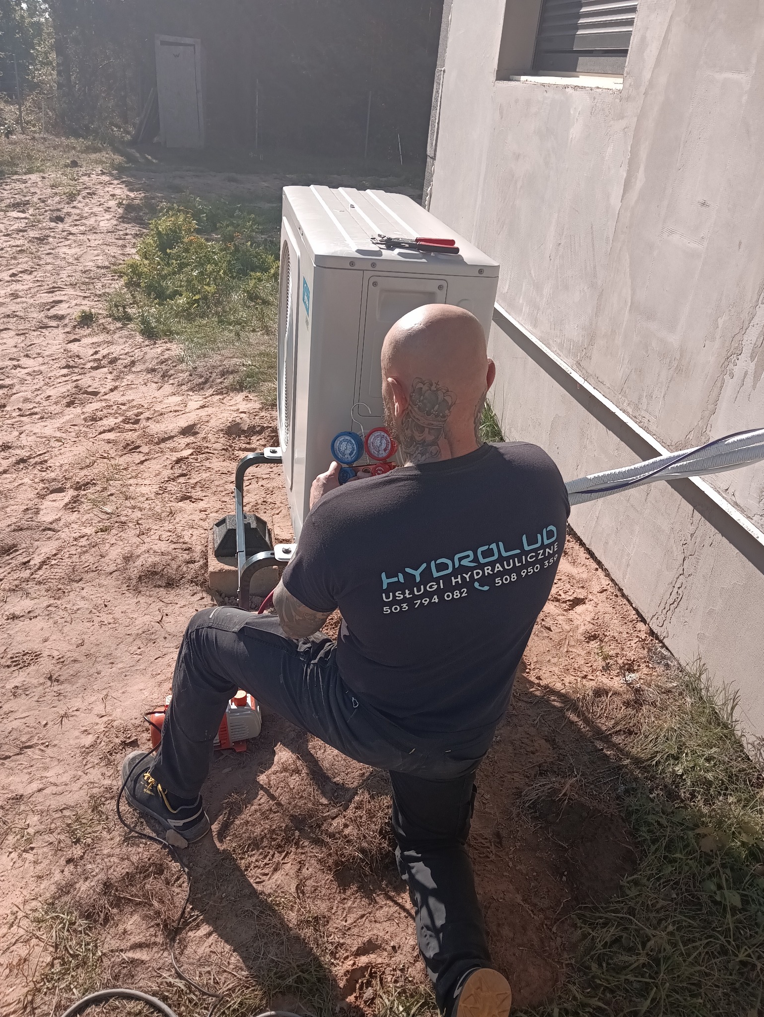 Hydraulik w trakcie montażu klimatyzacji zewnętrznej, używając manometrów. Na klimatyzatorze leżą narzędzia. Widoczny budynek w stanie surowym.