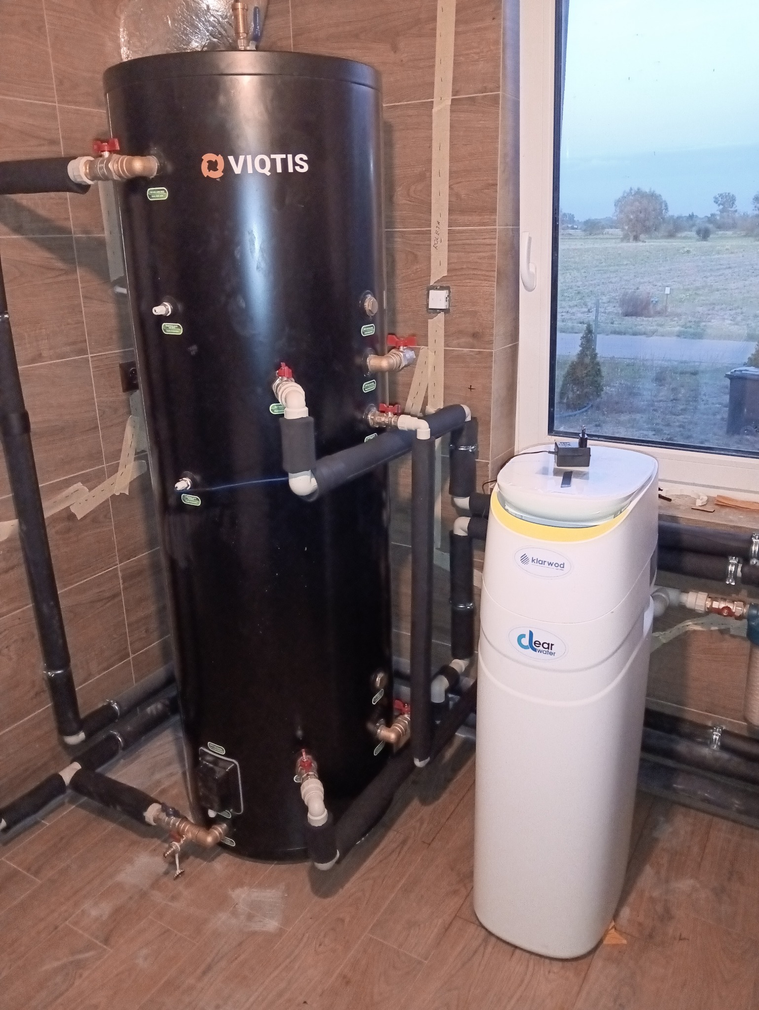 Czarny zasobnik ciepłej wody użytkowej Viqtis z podłączeniami i biały zmiękczacz wody Klarwod w pomieszczeniu z oknem i brązowymi płytkami.
