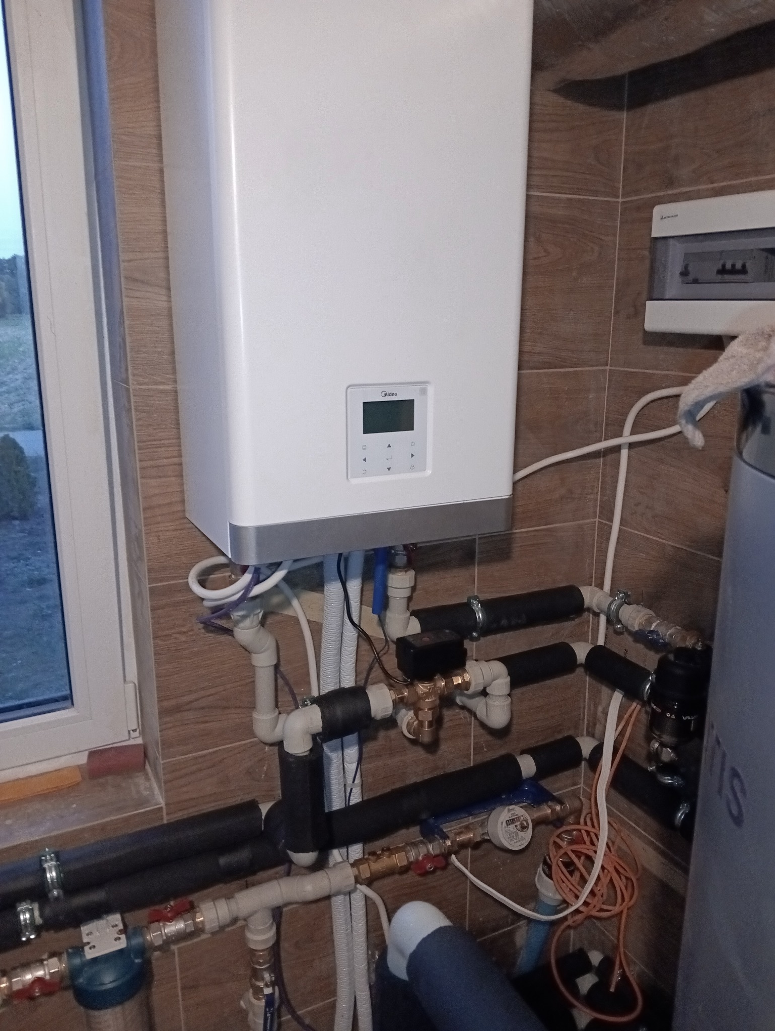 Biały piec gazowy Midea zamontowany na ścianie z brązowych płytek. Widoczne rury, zawory i filtr wody. Obok okno i szara obudowa elektryczna.