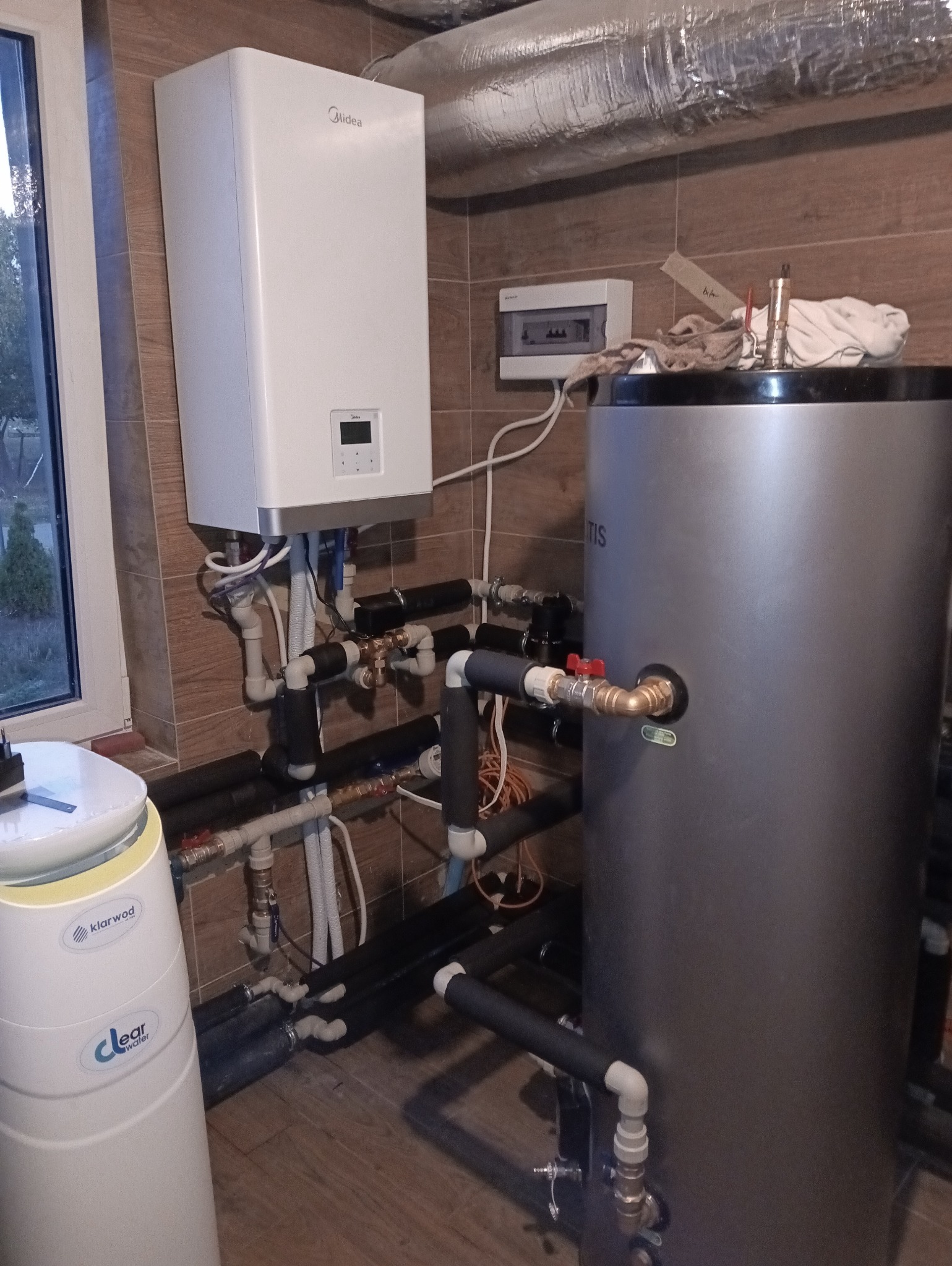 Biały piec gazowy Midea z szarą obudową bojlera, rury instalacyjne w izolacji, filtr wody Klarwod. Instalacja w brązowej płytkowej kotłowni.
