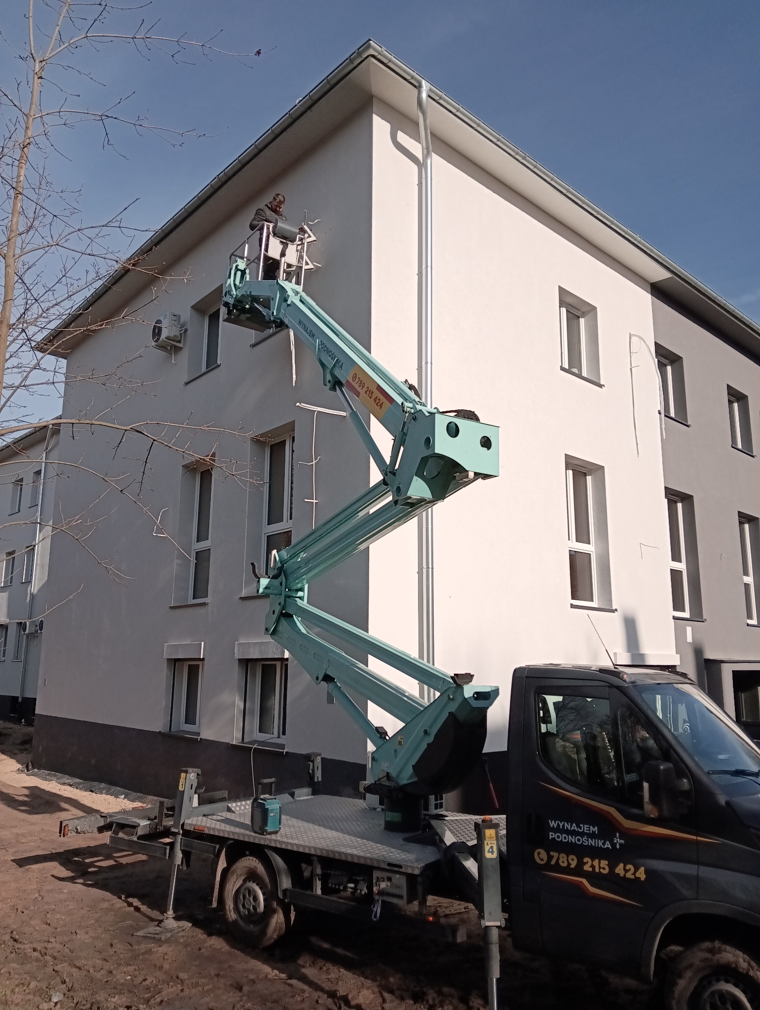 Praca na wysokości: pracownik w podnośniku koszowym przy elewacji budynku. Widoczny fragment elewacji, okna i klimatyzator. Na samochodzie numer telefonu.