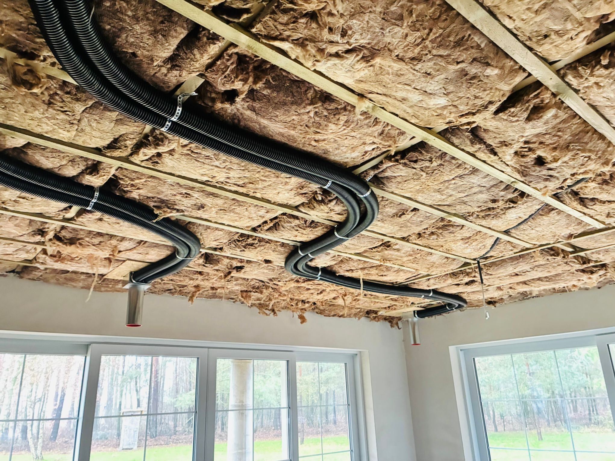 Izolacja stropu wełną mineralną z poprowadzonymi rurami wentylacyjnymi, widok z dołu na tle okna. Elementy instalacji przymocowane do drewnianej konstrukcji.