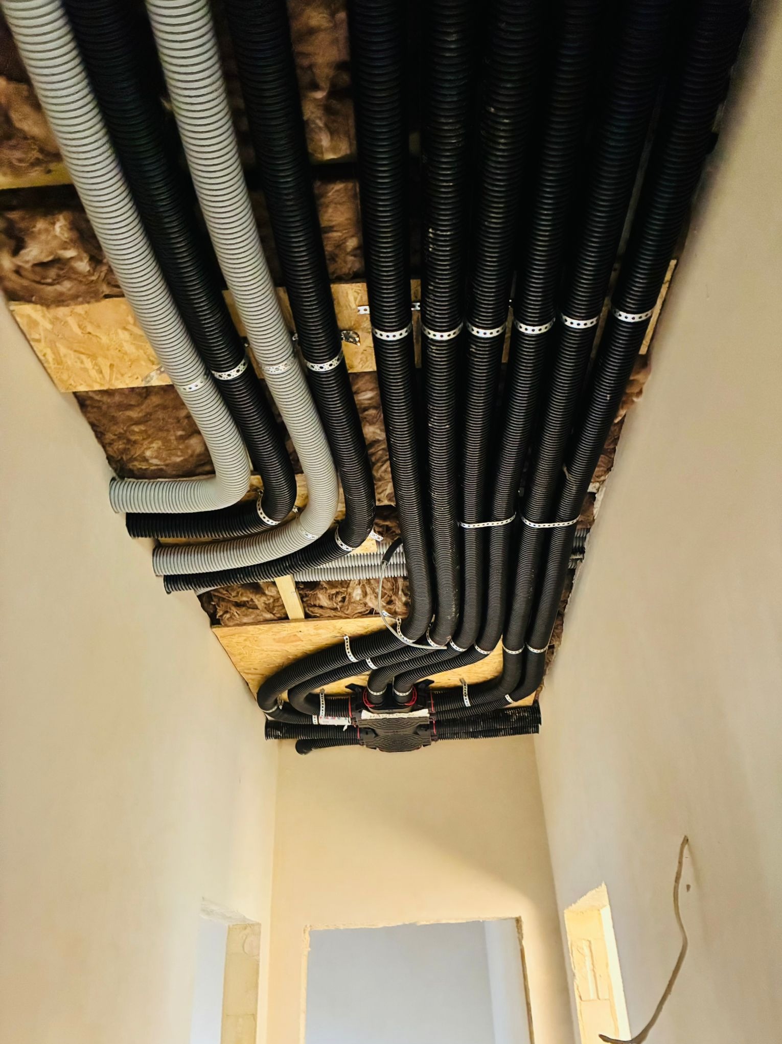 Równolegle ułożone, czarne i szare rury instalacyjne pod sufitem, przymocowane metalowymi obejmami. Widoczna izolacja i fragment ściany z otworem drzwiowym.