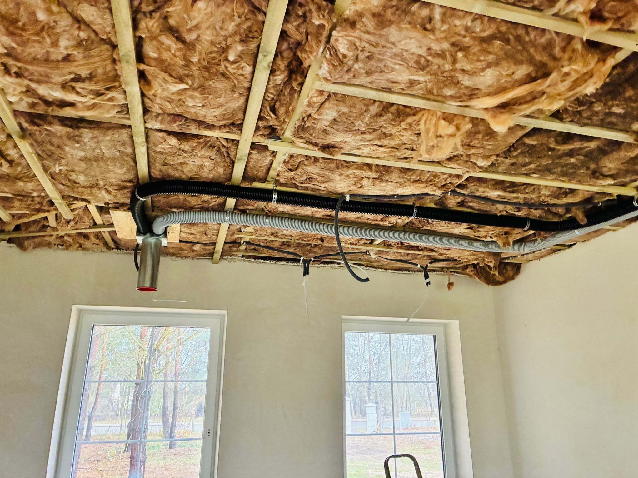 Ocieplenie stropu wełną mineralną z widocznymi przewodami wentylacyjnymi i elementami instalacji, w tle dwa okna. Prace wykończeniowe w nowym domu.