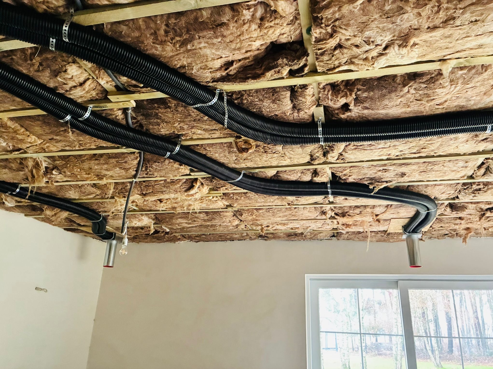 Czarna, karbowana rura instalacyjna przymocowana do sufitu z izolacją z wełny mineralnej, widok z dołu, fragment okna w tle.