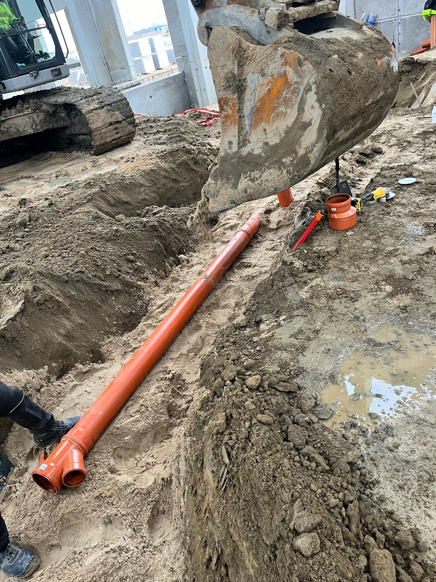 Pomarańczowa rura kanalizacyjna w wykopie, obok koparka i narzędzia. Widoczne elementy instalacji i etap prac ziemnych. Tło stanowią betonowe elementy konstrukcyjne.