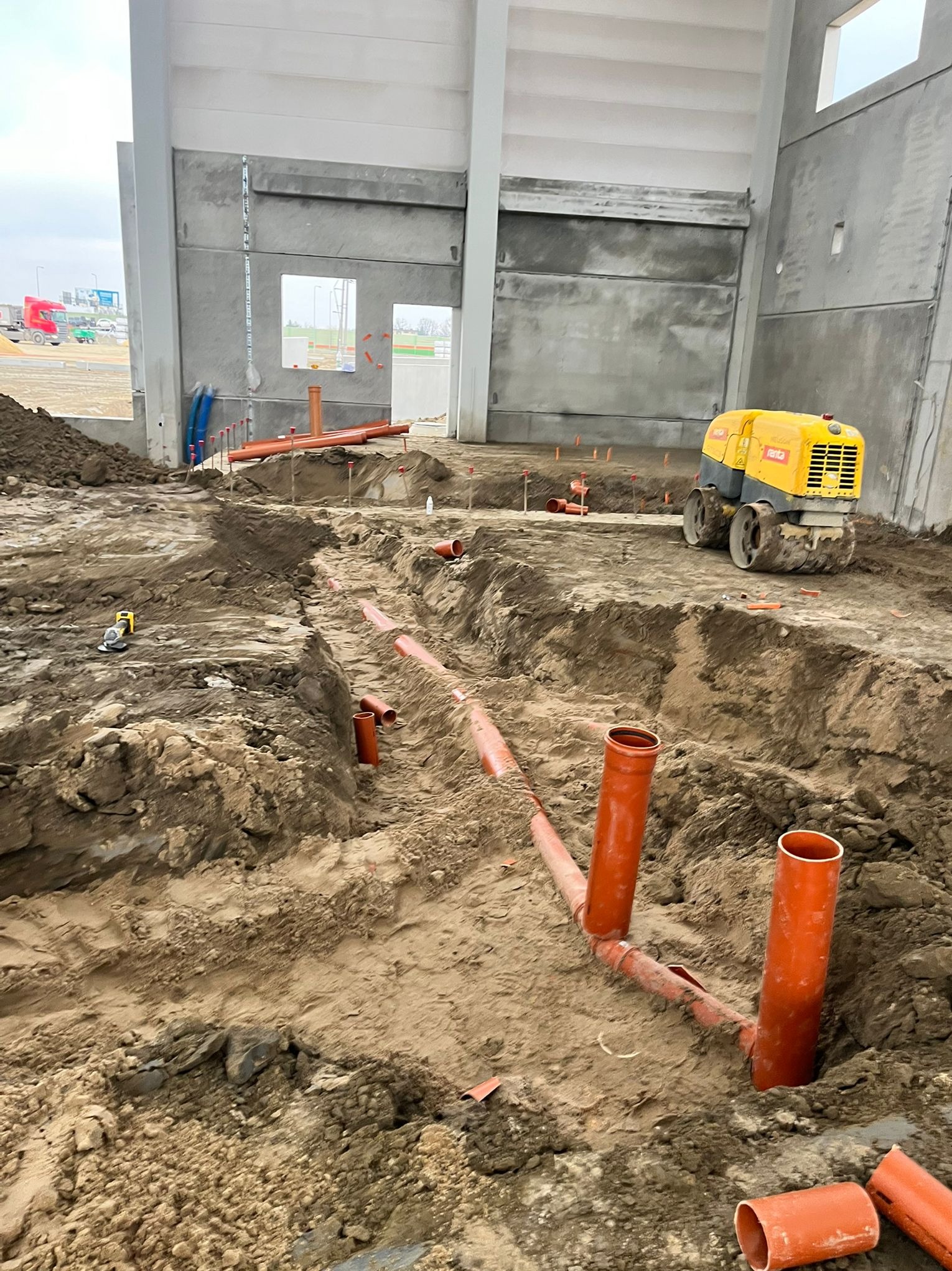 Pomarańczowe rury kanalizacyjne ułożone w wykopie ziemnym wewnątrz surowego budynku. Żółty walec wibracyjny w tle. Widoczne narzędzia i elementy instalacji.