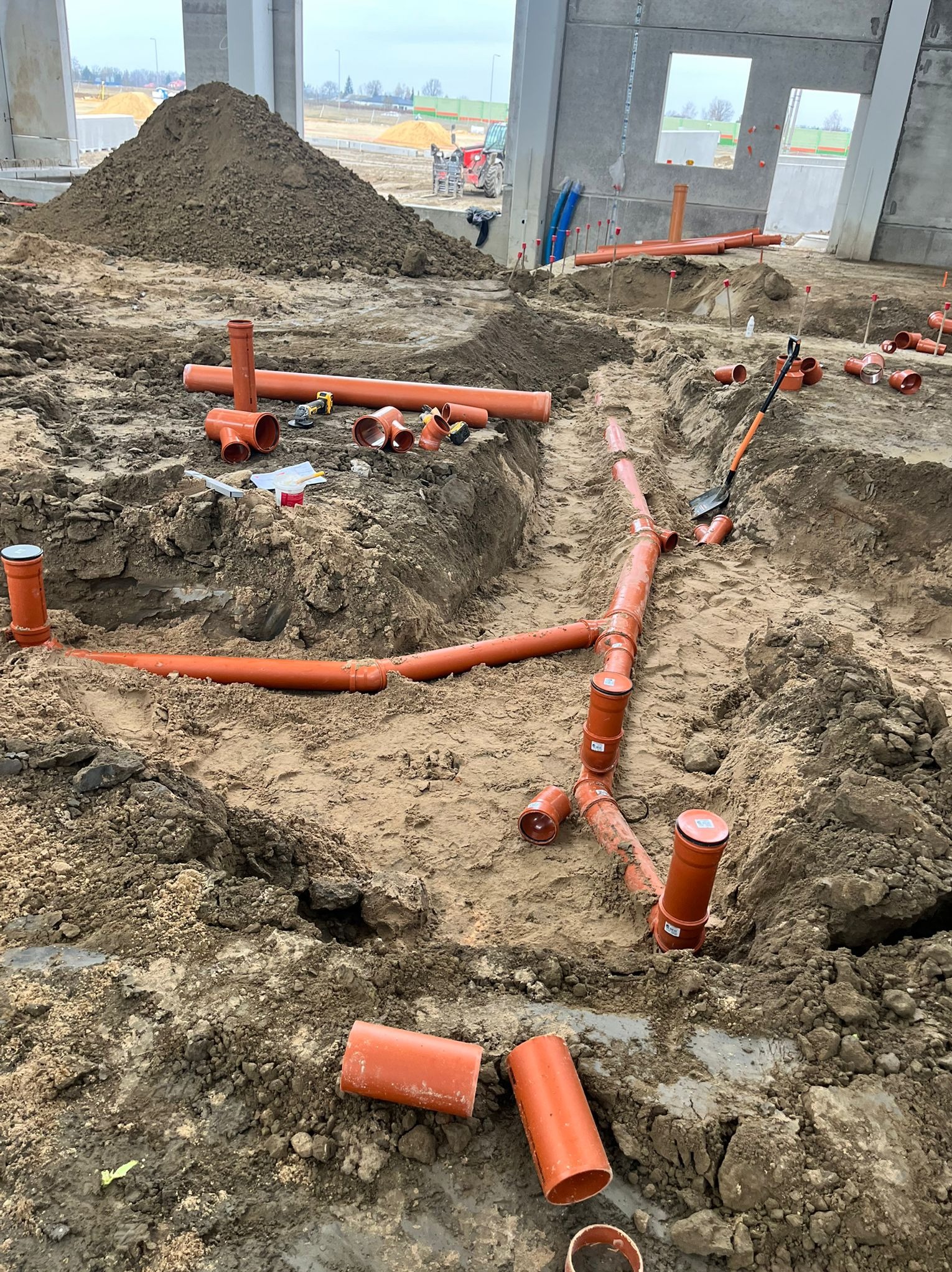Pomarańczowe rury kanalizacyjne ułożone w wykopie na placu budowy. Widoczne złączki, narzędzia i hałda ziemi w tle. Etap prac instalacyjnych w surowym stanie.