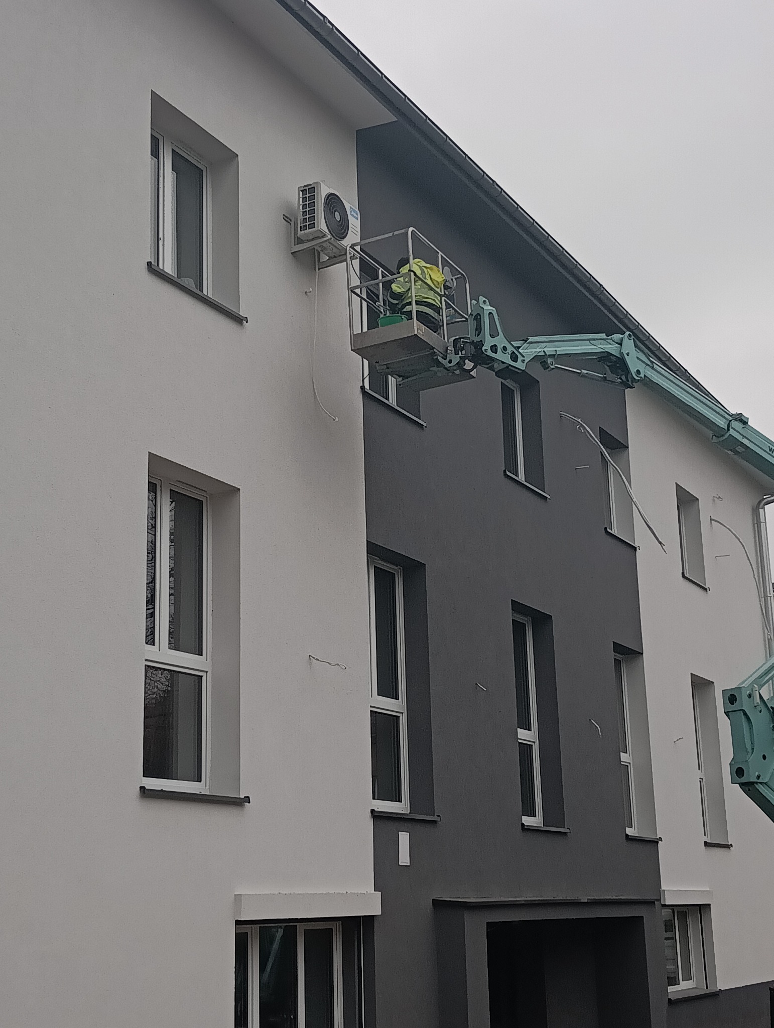 Montaż klimatyzacji na elewacji budynku z podnośnika koszowego. Widoczny pracownik w żółtej kamizelce i jednostka zewnętrzna klimatyzatora na ścianie.