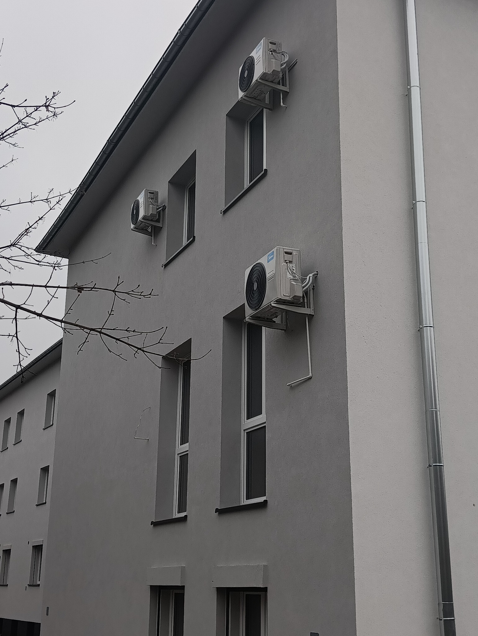 Szara fasada budynku z trzema zamontowanymi klimatyzatorami Midea, każdy z metalowym wspornikiem. Widoczne okna i rynna. Pochmurne niebo w tle.
