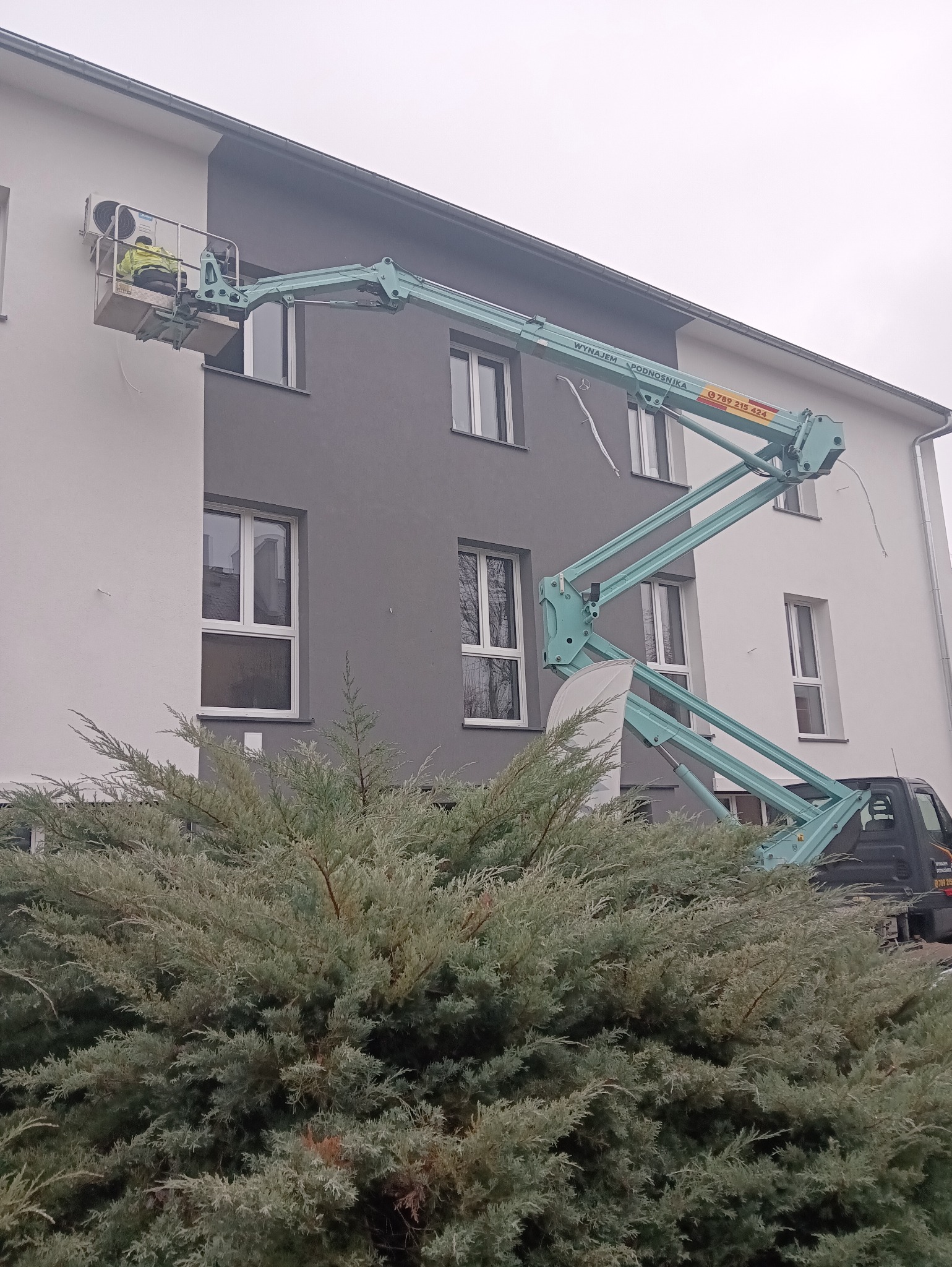 Montaż klimatyzacji na elewacji budynku z użyciem podnośnika koszowego. Pracownik w żółtej kamizelce w koszu podnośnika. Widoczny fragment krzewu na pierwszym planie.