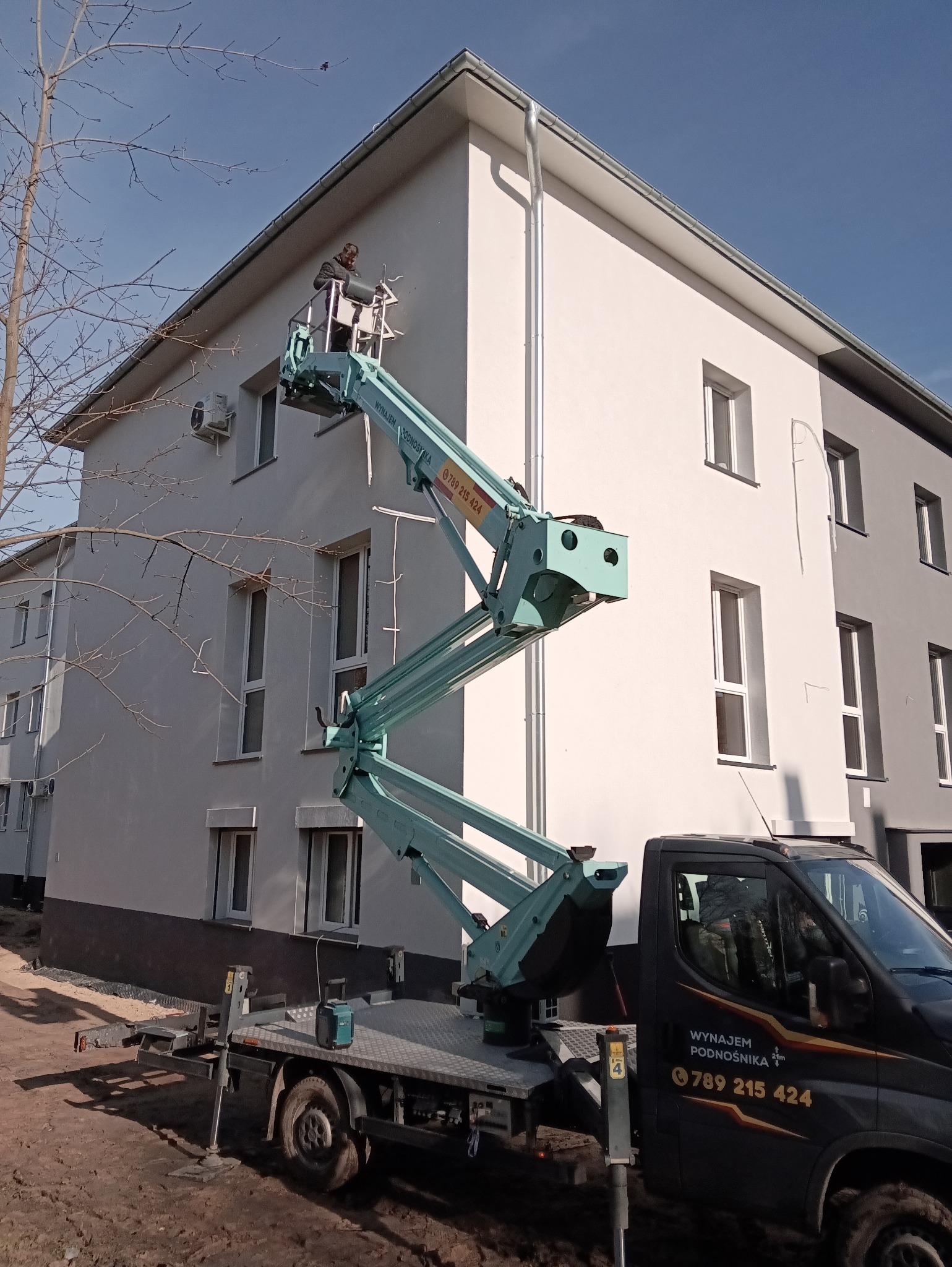 Pracownik na podnośniku koszowym montuje klimatyzację na elewacji budynku. Widoczny agregat klimatyzacyjny i fragment instalacji. Budynek dwukondygnacyjny, częściowo szary.