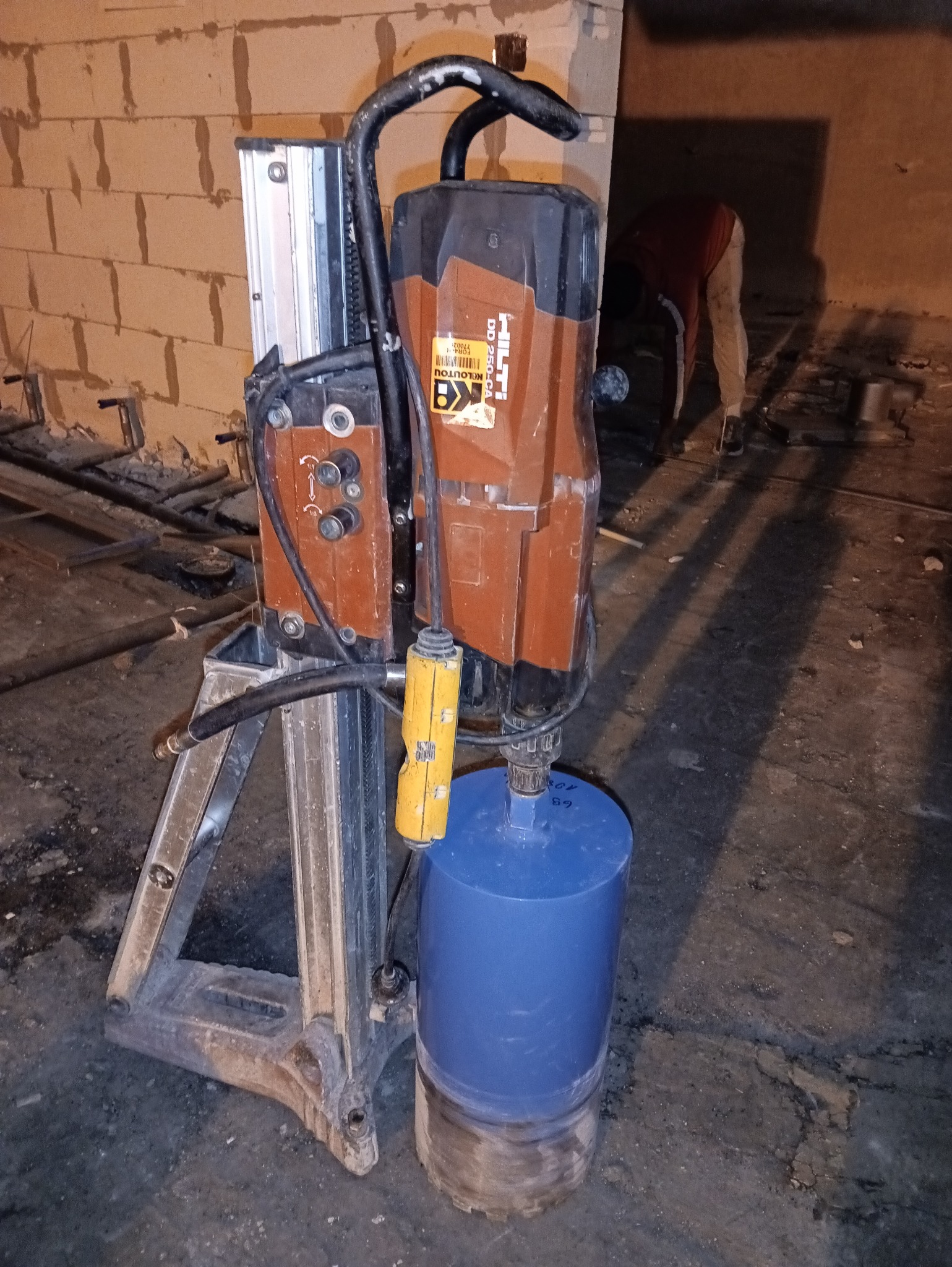 Wiertnica Hilti DD 250-CA z wiertłem koronowym na budowie, w tle mur z pustaków i osoba. Sprzęt do wiercenia otworów w betonie lub murze.