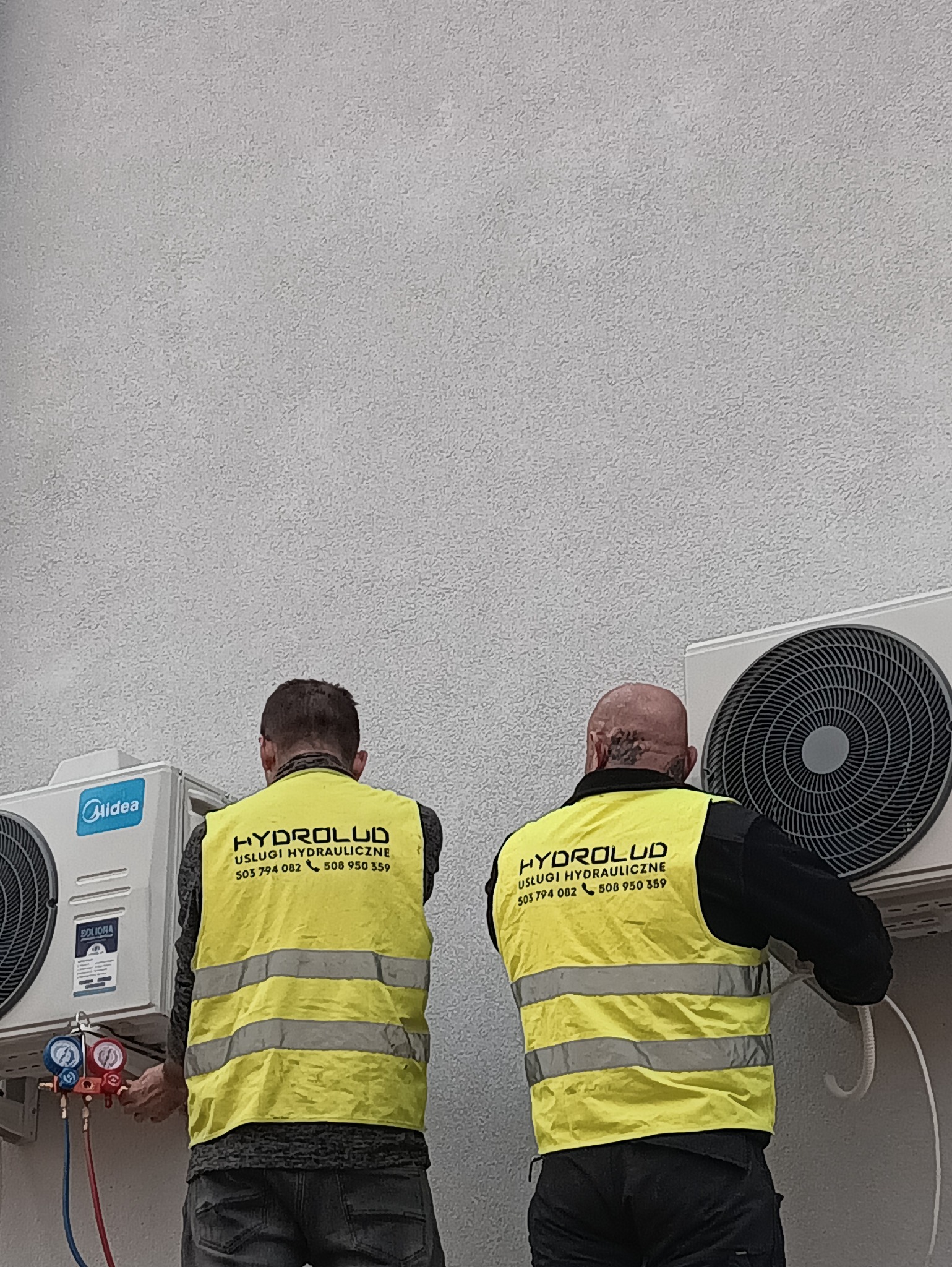Dwóch hydraulików w żółtych kamizelkach firmowych montuje klimatyzator Midea na jasnej ścianie. Widoczne narzędzia i manometry. Ujęcie od tyłu, z dołu.