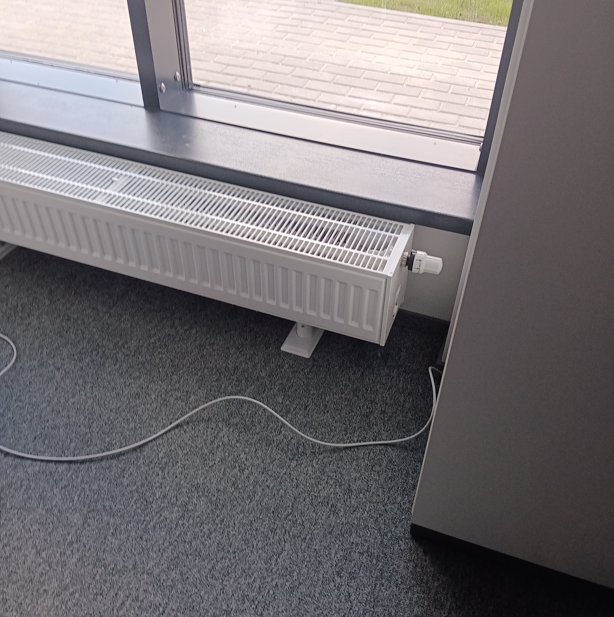 Biały, żebrowany grzejnik panelowy zamontowany pod oknem na szarym dywanie. Widoczny termostat i kabel. Ujęcie z niskiego kąta, z boku.