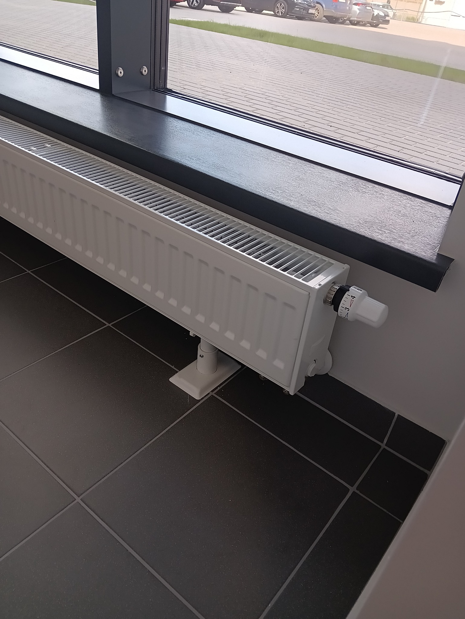 Biały grzejnik panelowy z termostatem, zamontowany pod oknem na ciemnej, kwadratowej płytce podłogowej. Widok z boku, z zewnątrz budynku w tle.