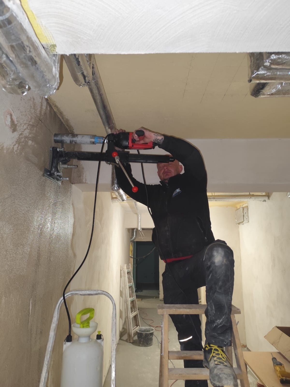 Pracownik na drabinie wierci otwór w ścianie za pomocą wiertnicy diamentowej. Widoczne rury pod sufitem i ściana w trakcie prac wykończeniowych. W tle korytarz.
