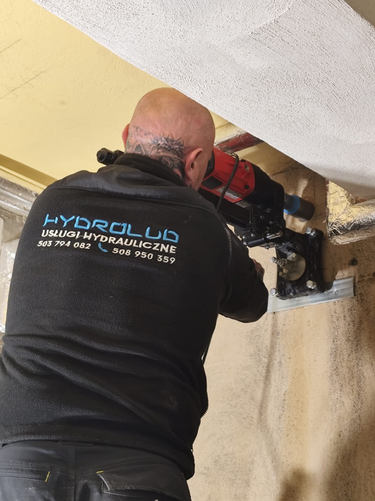 Hydraulik wierci otwór w ścianie, używając wiertnicy diamentowej. Widoczny fragment instalacji i logo firmy na bluzie pracownika.